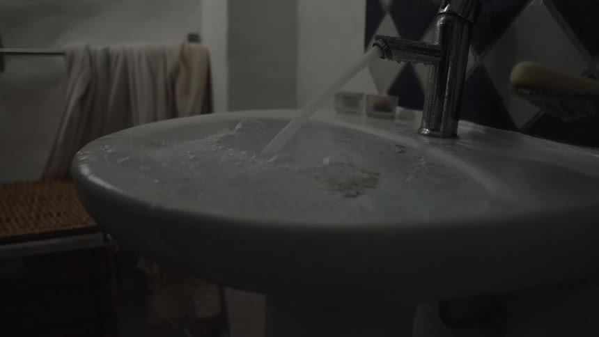 Running faucet filling a white sink in a bathroom with dark tiles