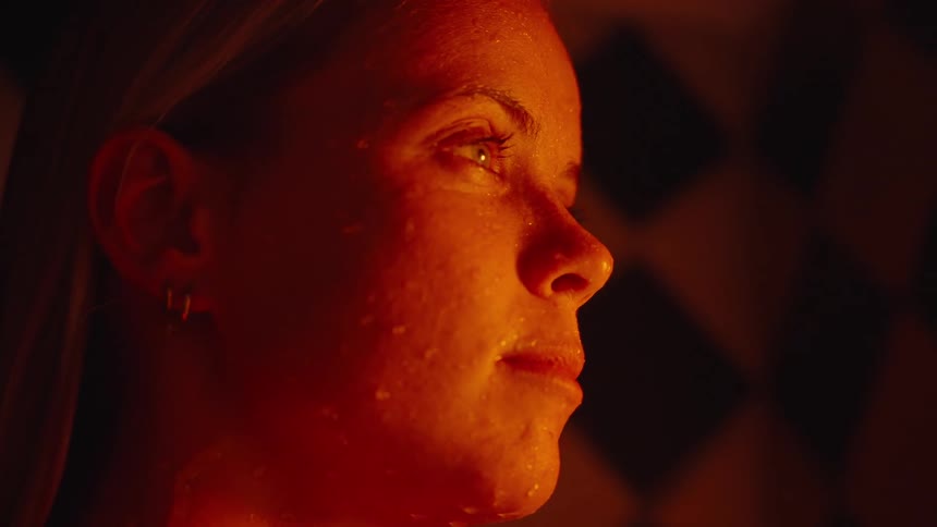 The video is a closeup shot of a persons upper body and face being drenched by warm water possibly in a sauna or a dimly lit shower the skin is glowing orangered from the lighting and the water runs down the face and neck with a thin gold n