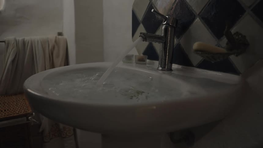 Water pours from a shiny metal faucet into a white bathroom sink which quickly fills and overflows with a hand briefly touching the water before it stops flowing in a dimly lit room with blue and white tiled walls