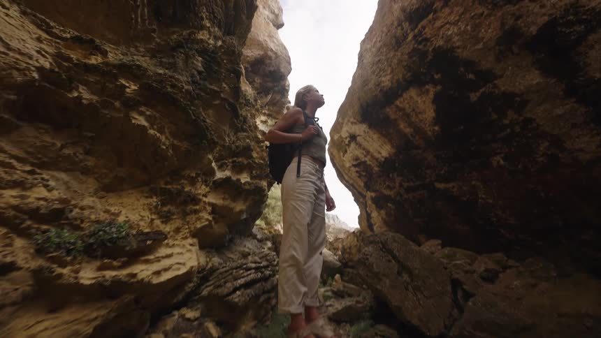 A low angle shot shows a woman wearing a tank top tan pants and a black backpack standing on a large rock looking up at the bright sky between two tall golden brown rock formations in a sunlit canyon