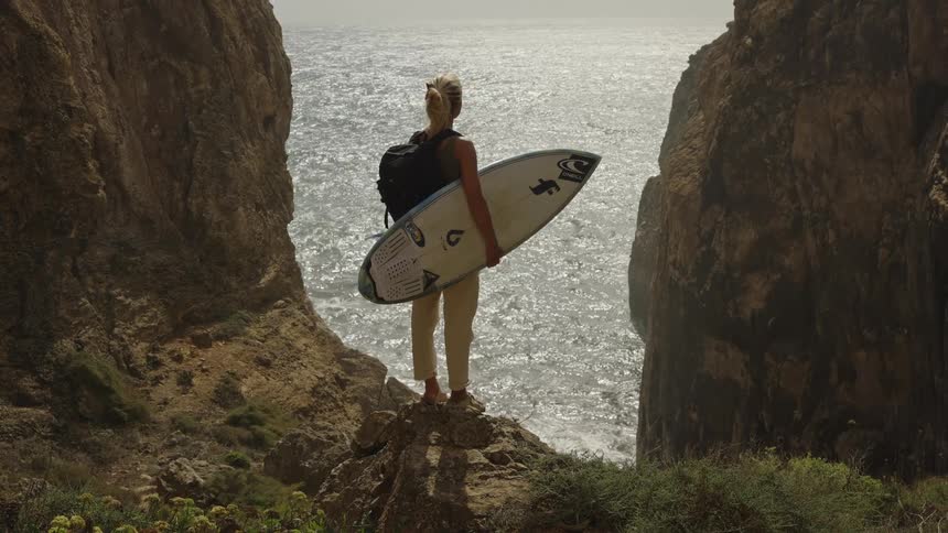 A young blonde woman wearing a black backpack and tan pants stands on a rocky cliff edge holding a white surfboard looking out at the bright sunlit ocean between two massive rock walls with a white dog visible in the foreground