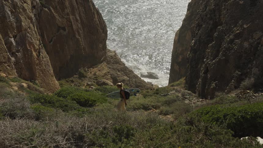 A high wide view shows a blonde woman carrying a surfboard and walking downward on a steep brush covered hillside between massive sunny rock cliffs toward the glittering ocean far below
