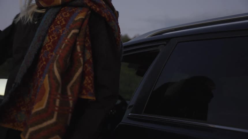 A side view shows a blonde woman wearing a colorful patterned scarf getting out of a dark suv closing the driver side door on a dull overcast day with dark foliage in the background
