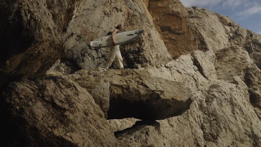 A young woman carries a white surfboard steps and walks across large uneven rocks tall rock cliffs surround the person and the setting is outdoors under sunlight