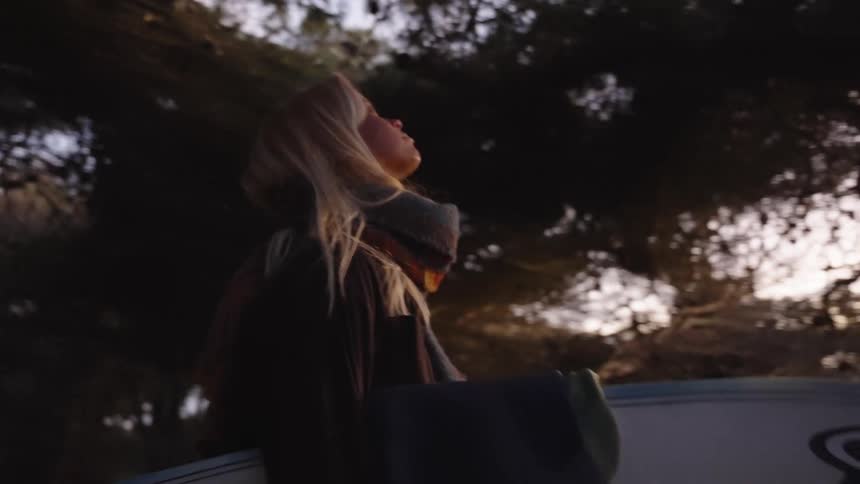 A close backlit view shows a blonde woman wearing a dark jacket and a patterned scarf carrying a surfboard on her hip looking upward with the wind blowing her hair among dark pine trees at dusk