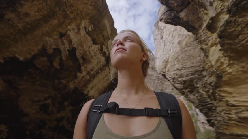 A low angle close view shows a blonde woman wearing a green tank top and a black backpack standing in a narrow rock gorge looking upward at the bright sky and the massive rugged golden brown walls