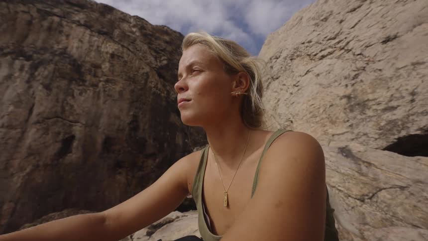 A young blonde woman wearing a green tank top and a necklace is shown in a close outdoor shot turning her head and looking around a rocky sunlit gorge