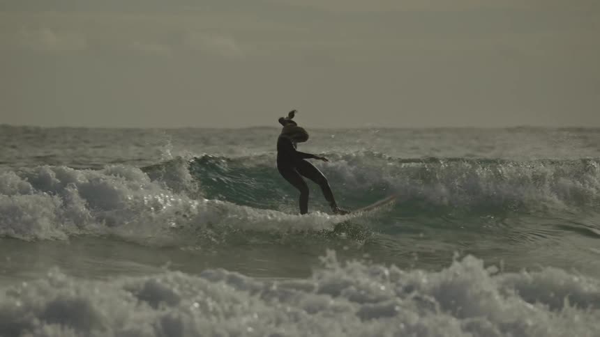 A woman surfs a wave near a cliff under a bright overcast sky