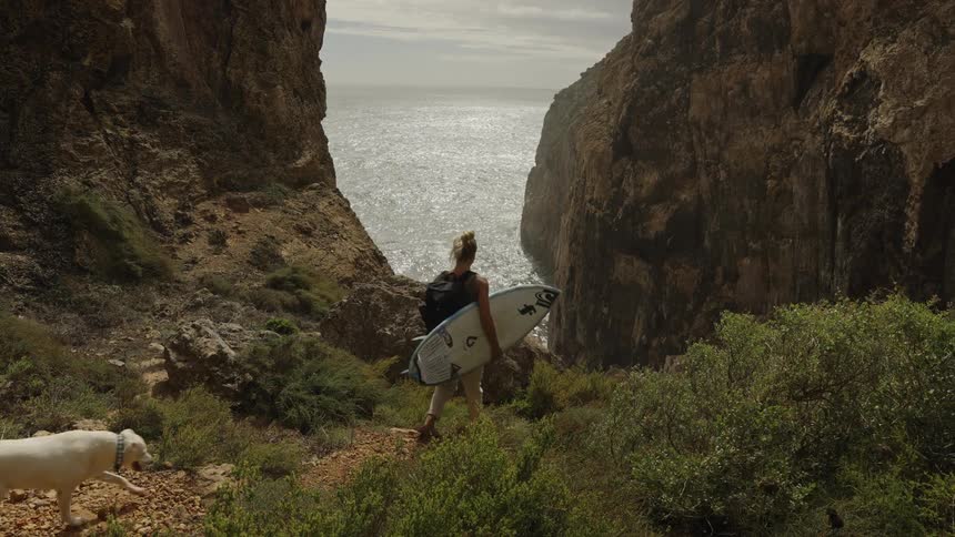 A person with a surfboard and a backpack walks down a dirt path a dog follows the person tall rock cliffs frame a view of the ocean the water surface reflects sunlight and scrub vegetation covers the ground