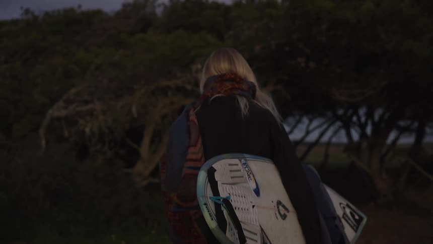 A medium shot from behind shows a blonde woman wearing a dark jacket and a patterned scarf walking away from the camera through dark dense trees carrying a white surfboard under her arm at dusk