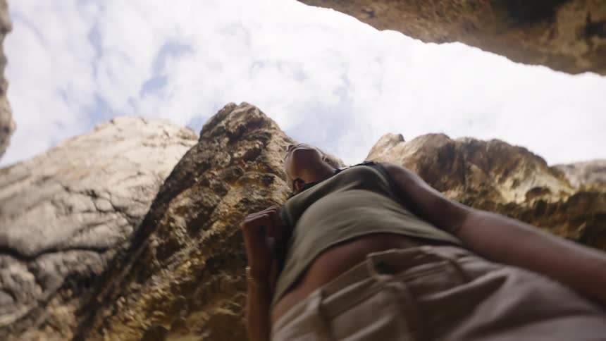 A unique lowangle view looking upward shows a woman wearing a green shirt and tan pants standing between two tall rough rock walls in a natural canyon with bright sky visible above