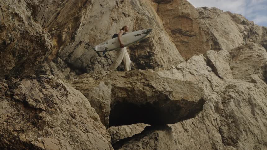 Person carrying a surfboard climbing rocky formations near the sea