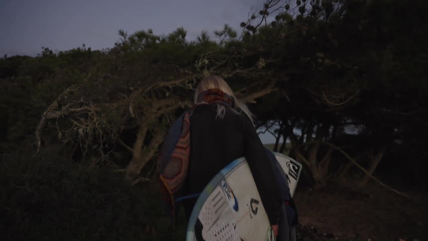 Woman walks along a wooded trail carrying a surfboard at dusk