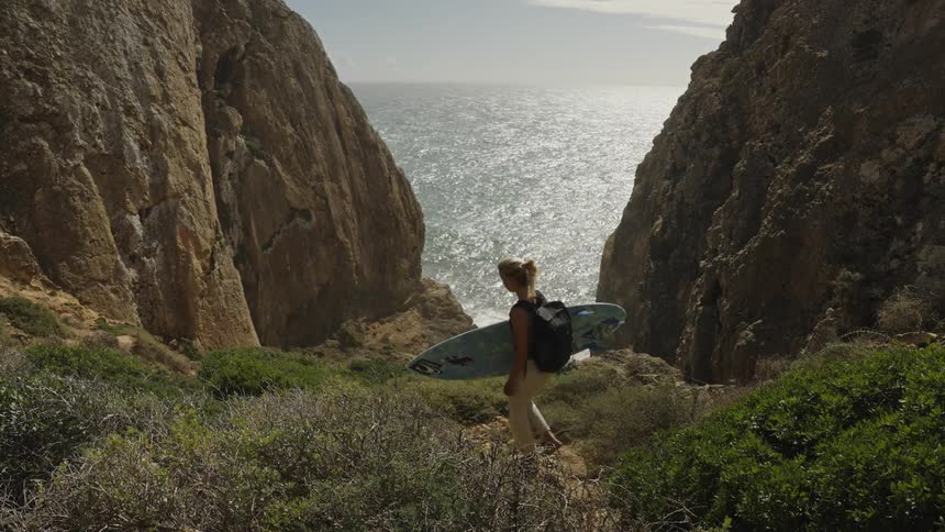 Woman carrying a surfboard toward the sea between cliffs