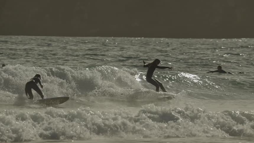 The video is an extreme closeup shot of two surfers riding a small wave towards the shore framed against the dark ocean and a distant large cliff both surfers are silhouetted in dark wetsuits as they ride the greenandwhite crest of the wave