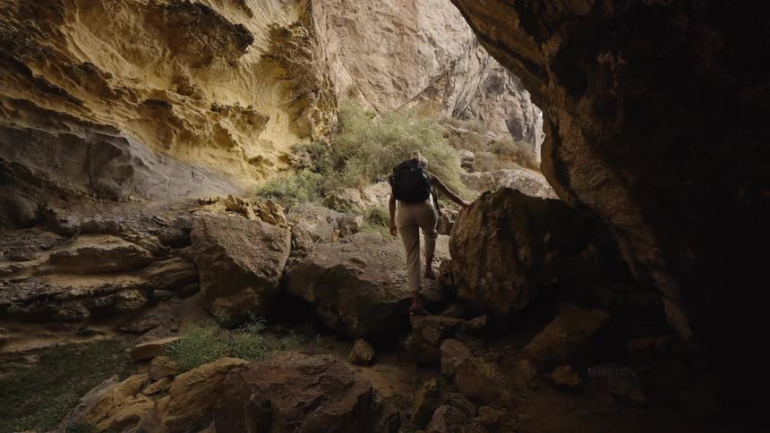 Woman with a backpack climbs among rocks on a trail inside a sunlit canyon