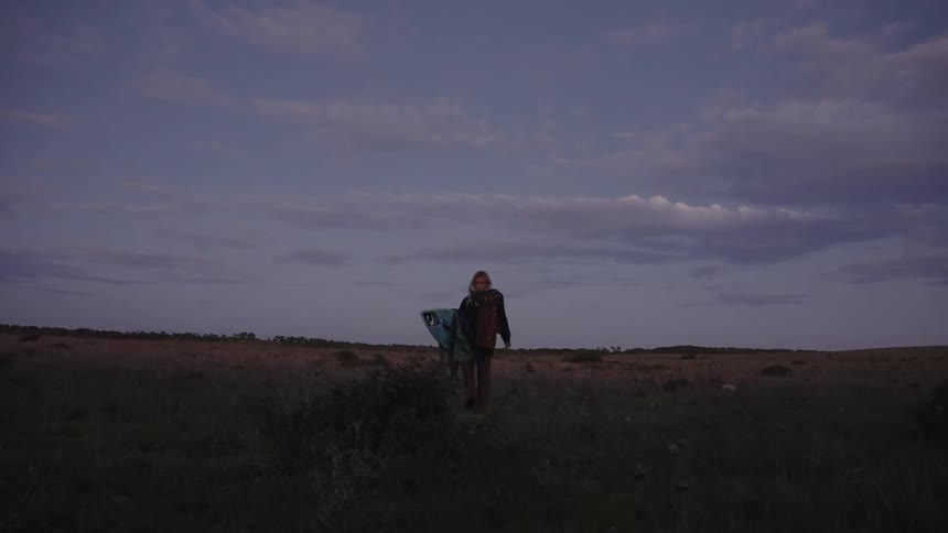 Woman walking across an open field at dusk carrying a surfboard