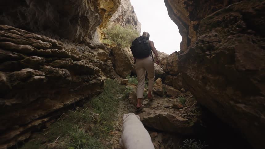 A view from behind a white dog shows a woman wearing a black backpack and light colored pants climbing over large rocks and up a narrow dark canyon path