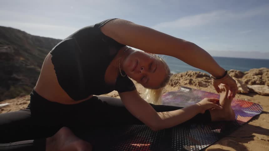 A close low angle shot shows a blonde woman in a black crop top and leggings performing a seated side bend yoga stretch on a colorful mat on a sunny dusty cliff overlooking the ocean