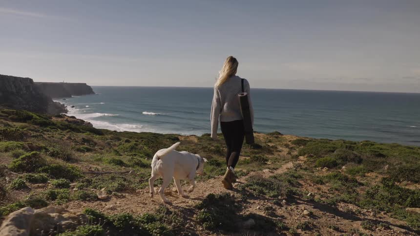 A person walks down a dirt path on a hillside a white dog follows close behind the person the person carries a rolled mat on the shoulder the ocean and shoreline cliffs stretch across the background under bright sun