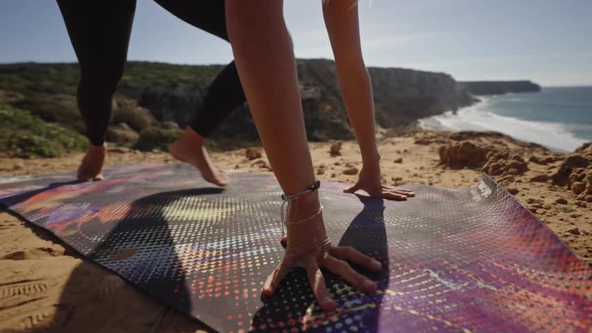 A person performs a yoga pose on a mat the person moves from a lunge to a downward facing pose the yoga mat rests on the ground hands and feet touch the mat and the background shows the ocean and a cliff
