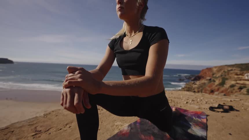 A blonde woman wearing a black crop top and leggings is shown in a close view doing a lunge stretch on a patterned mat on a sandy cliff edge with a beach and ocean visible in the background on a sunny day