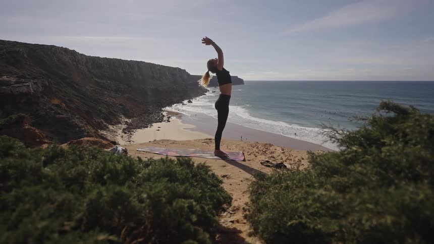 Blonde woman performs yoga on a small beach while her dog walks nearby