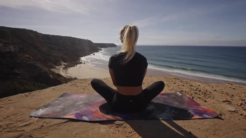 The video shows a woman sitting crosslegged on a yoga mat on a clifftop looking out over a beach and the blue ocean the scene is bright and sunny featuring steep eroded cliffs plunging down to the sandy shore