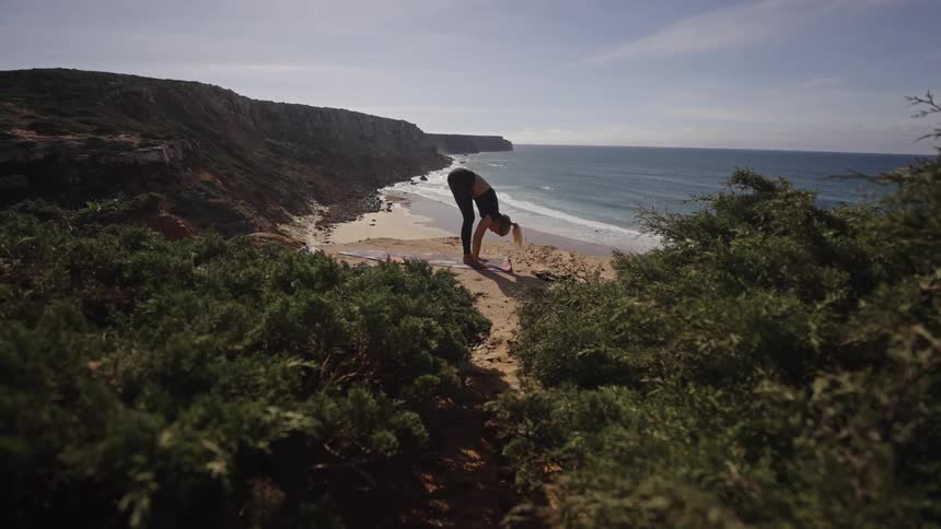 A blonde woman wearing black workout clothes is seen performing yoga stretches on a mat on a sandy path on a high cliff overlooking a secluded beach and sunny ocean