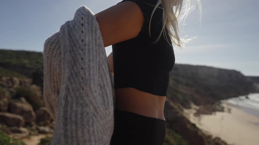A blonde woman is shown in a close view on a cliff top on a sunny day taking off a thick grey knitted cardigan to reveal a black sports bra and leggings with a secluded beach and ocean visible far below