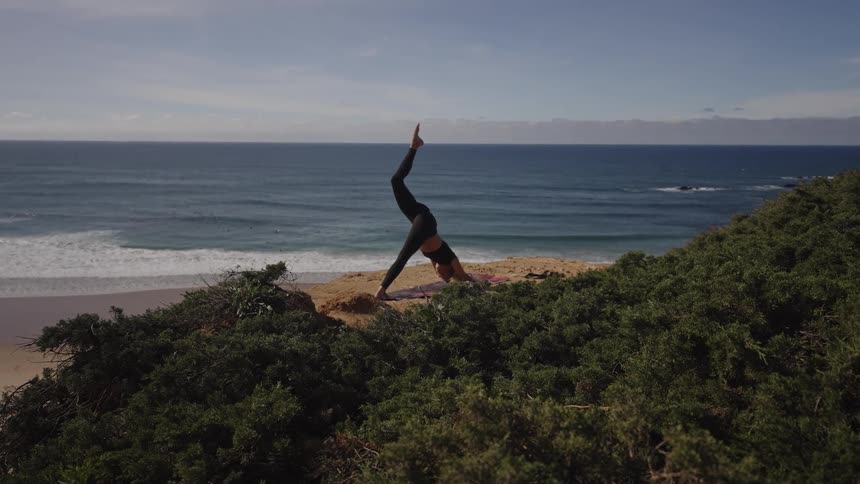 Woman practicing a yoga pose with one leg raised facing the ocean