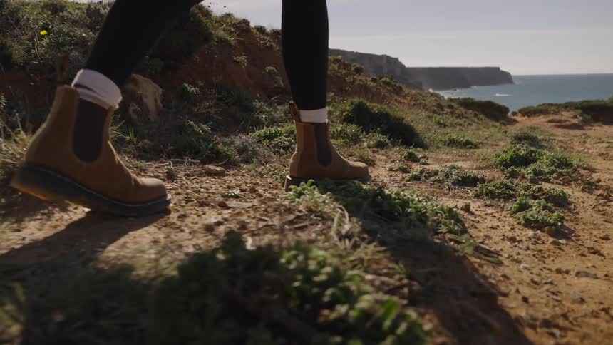 The video is a closeup of a persons brown boots walking on a dusty rocky coastal cliffside trail the ocean is visible in the background along the horizon under a bright sky