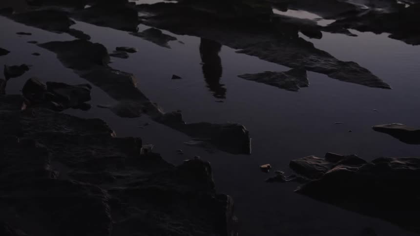 A shallow pool of water rests among dark rocks the water reflects a blurred dark outline of a person the persons reflection moves slightly and the scene is outdoors in very low light