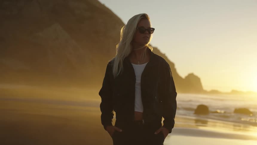 A young blonde woman wearing sunglasses and a jacket standing and looking to the side at the beach