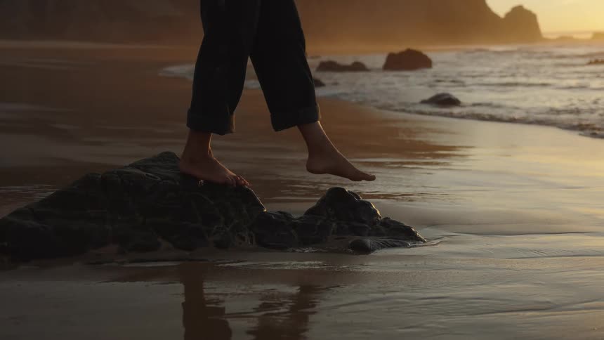A person with bare feet and rolled up dark pants steps off a dark rock onto the wet sandy beach as the sun sets in the background