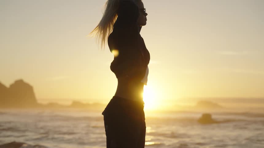 A person stands facing sideways on the shore the person raises their arms toward their head the sun sets over the ocean directly behind the person cliffs are visible in the distance and the persons body is mostly shadowed