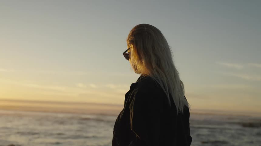 A beautiful blonde woman wearing sunglasses and a dark jacket over a white shirt stands on a sandy beach looking out at the ocean during sunset followed by a close view of her bare feet walking through the shallow water