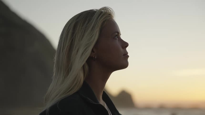 A young blonde stands on the beach sand at sunset feeling the warmth of the air