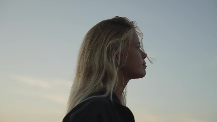 A person with long blonde hair looks upward in profile against a light sky the persons head is tilted back her hand briefly touches the forehead and she is walking in an outdoor setting