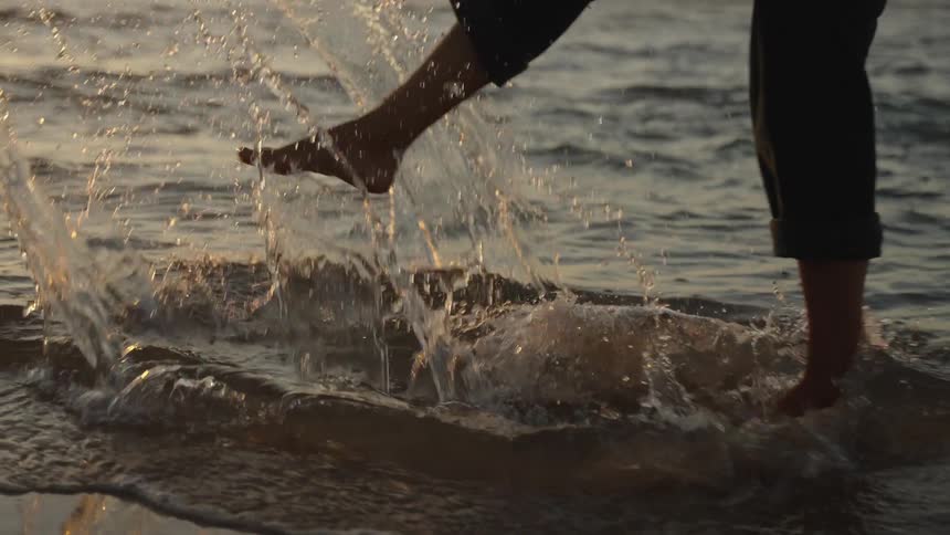 A person stands barefoot in shallow ocean water near the shore one foot quickly kicks the water upward creating a large sunlit spray and the golden light of sunset reflects off the waters surface