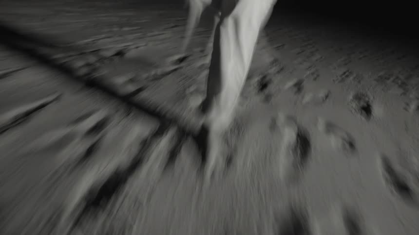 A black and white low angle view shows the legs of a person wearing light colored pants walking quickly across dark sand or dirt with the motion creating a blurry dynamic effect