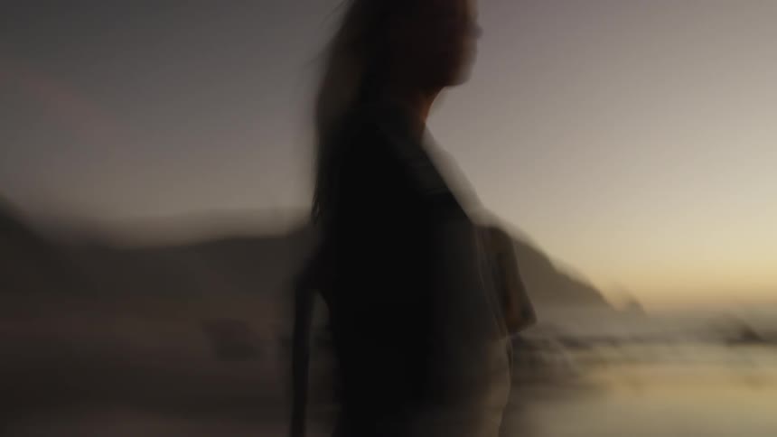 The video shows a blurry silhouetted figure of a woman with long light hair standing on a beach at dusk or dawn the light source is behind her casting the figure in shadow against the bright horizon and blurred landscape