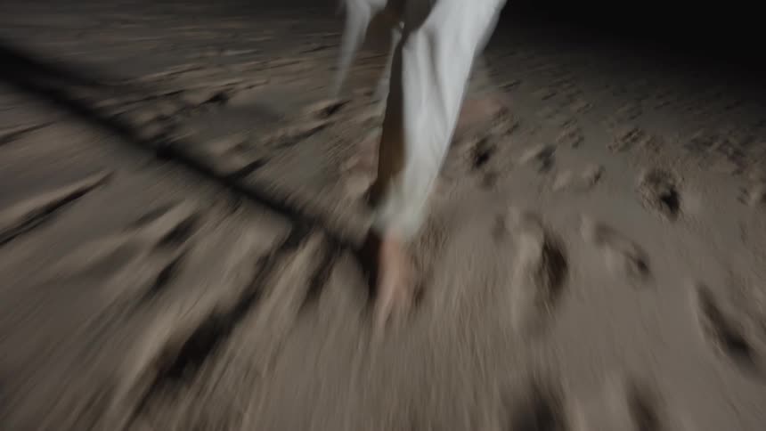 The video is an extreme closeup shot of a persons bare feet and lower legs running on sandy ground in the dark the motion blur and low light create a sense of urgency and frantic movement