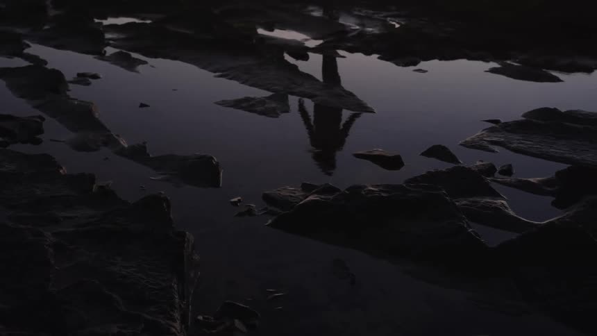 A shallow pool of dark water sits among jagged rock formations the surface reflects the inverted image of a persons legs and feet and the surrounding environment is characterized by dark colors and low light