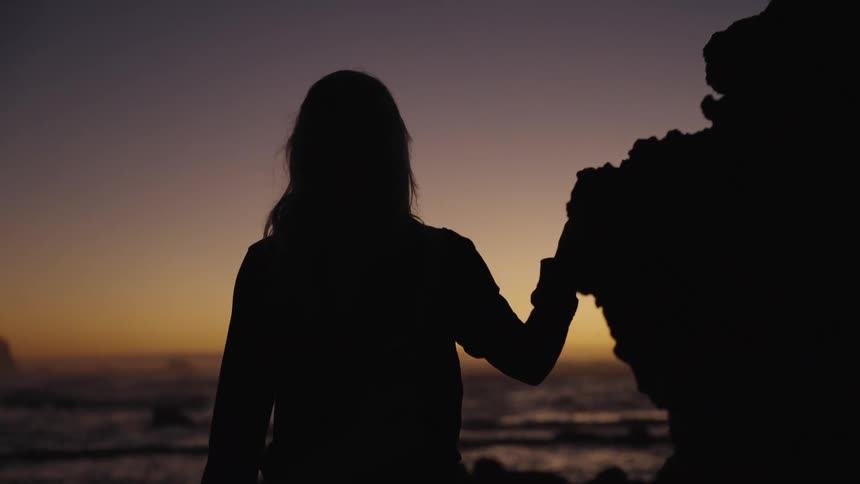 A low angle medium view shows the dark silhouette of a woman with long hair wearing a jacket and pants as she touches a tall rock outcrop and walks toward the dark ocean at sunset