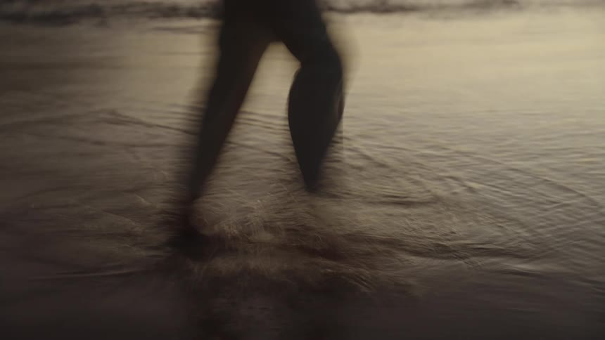 A dark motion blurred view focuses on the legs of a person wearing light colored pants walking barefoot through shallow shimmering water on a beach at dusk