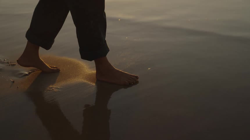 A persons bare feet walk across wet sand at the waters edge
