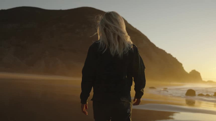 Blonde woman wearing sunglasses and a fashionable outfit walks on the beach while the sun shines on the horizon