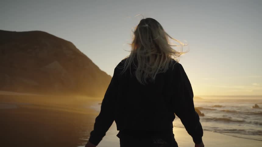 A person walks slowly along the wet sand of a beach the sun sets over the ocean surface creating a bright backlight large dark cliffs rise up on the left and the person turns their head from facing forward to looking out at the water