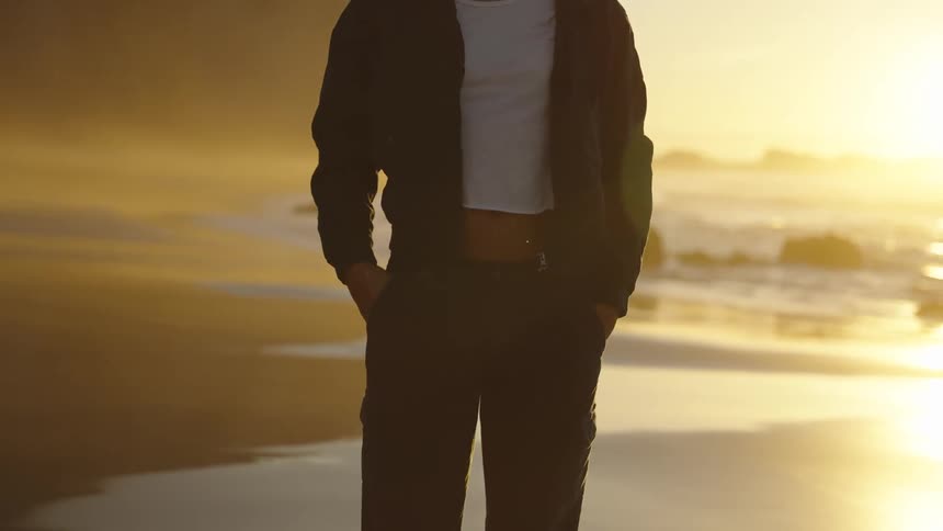 A blonde woman wearing sunglasses and a dark jacket over a white shirt stands on a wet sandy beach in the bright warm sunset light looking at the camera with steep rocky cliffs in the background